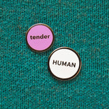 Round enamel pin that says TENDER next to a slightly larger white enamel pin that says HUMAN.  They are on a teal knitted background.