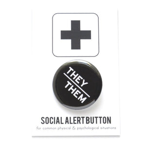 Round pinback button with black background which reads THEY/THEM  on an angle in white text, in a san serif font. On a white backing card, black plus sign at the top. Below reads: Social Alert Button, with tiny text beneath it which says, for common physical & psychological situations