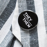 Round pinback button with black background which reads THEY/THEM  on an angle in white text, in a san serif font. Pronoun pin is pinned on a white & gray textile blazer lapel