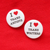 Two round white pinback buttons that reads I LOVE TRANS ARTISTS with love being represented by a red heart. Pinned to a red fabric background