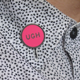 Round, neon pink enamel pin that says UGH on the collar of a polka dot shirt