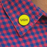 Neon yellow round enamel pin the says UGH on the collar of a pink and blue gingham shirt.