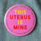 Big 3-inch shiny deep pink pinback button with bright yellow and red chunky san serif text that reads: This Uterus is Mine. Smaller text in a green runs in a circle around the permitter that says Fuck Off Alito, Fuck Off Thomas, Fuck Off Coney-Barrett, Fuck Off Gorsuch, Fuck Off Kavanaugh. The button is pinned to a worn denim jacket.