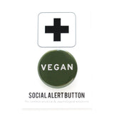 A round dark green (the color of leafy Dino kale) pinback button that reads VEGAN in white text. Badge is on a Social alert Button backingc card