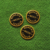 Three round WOMEN'S WEED LEAGUE enamel pins on a green textile background.