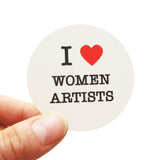 Round die-cut sticker that says I LOVE WOMEN ARTISTS. The text is black with a red heart signifies the word love. The background is white.