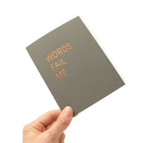Dark gray vertical greeting card with a thin san serif font stamped in copper hot foil , reading WORDS FAIL ME. The card is held up by a hand from the left hand corner, at an angle, on a white background