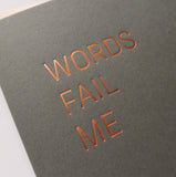Close up of copper hot foil text on a dark gray greeting card with a thin san serif font stamped in copper hot foil , reading WORDS FAIL ME