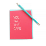 Vertical rectangle greeting card in a rich vibrant coral color with fun glitter hot foil embossed to say YOU TAKE THE CAKE. Pictured with coordinating aqua envelope. Aqua tous les jours pen rests diagonal across the upper right corner.