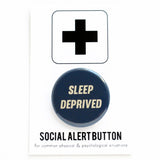 Round navy blue pinback button that reads SLEEP DEPRIVED in cream slanted text. Pinned to a Social Alert Button backing card.