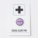 Round enamel pin that says TENDER on a Social Alert Pin backing card.